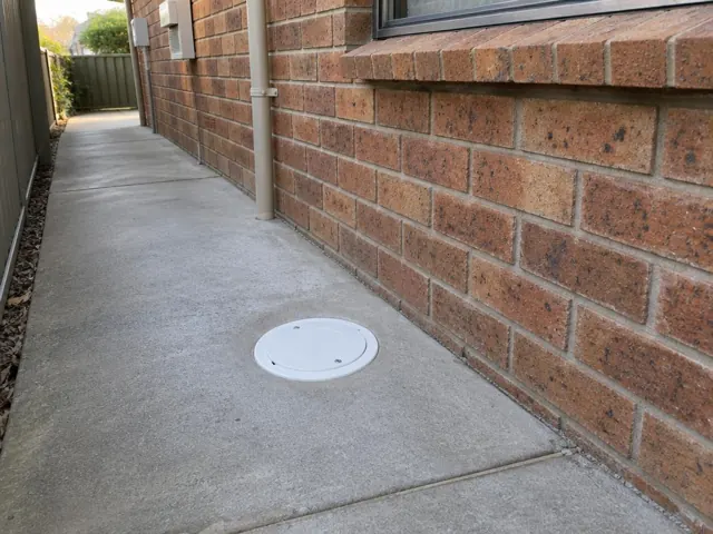 Capped round inspection opening in a concrete side path beside a brick house in Greystanes