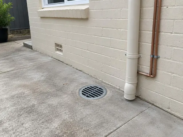 Outdoor gully grate set in concrete beside cream brick wall and downpipe in Wentworthville